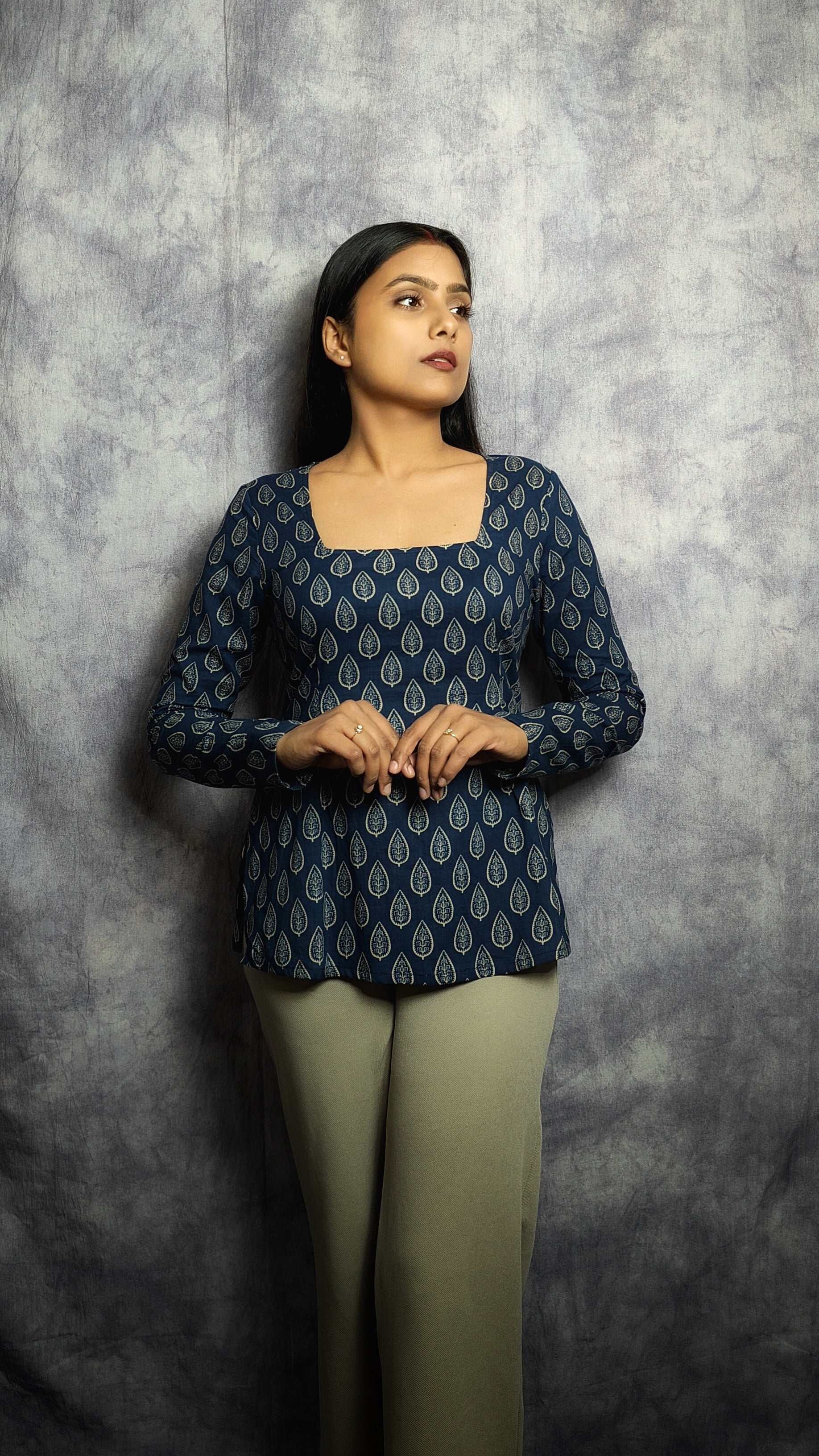 Woman wearing a dark blue patterned top and green pants against a gray background